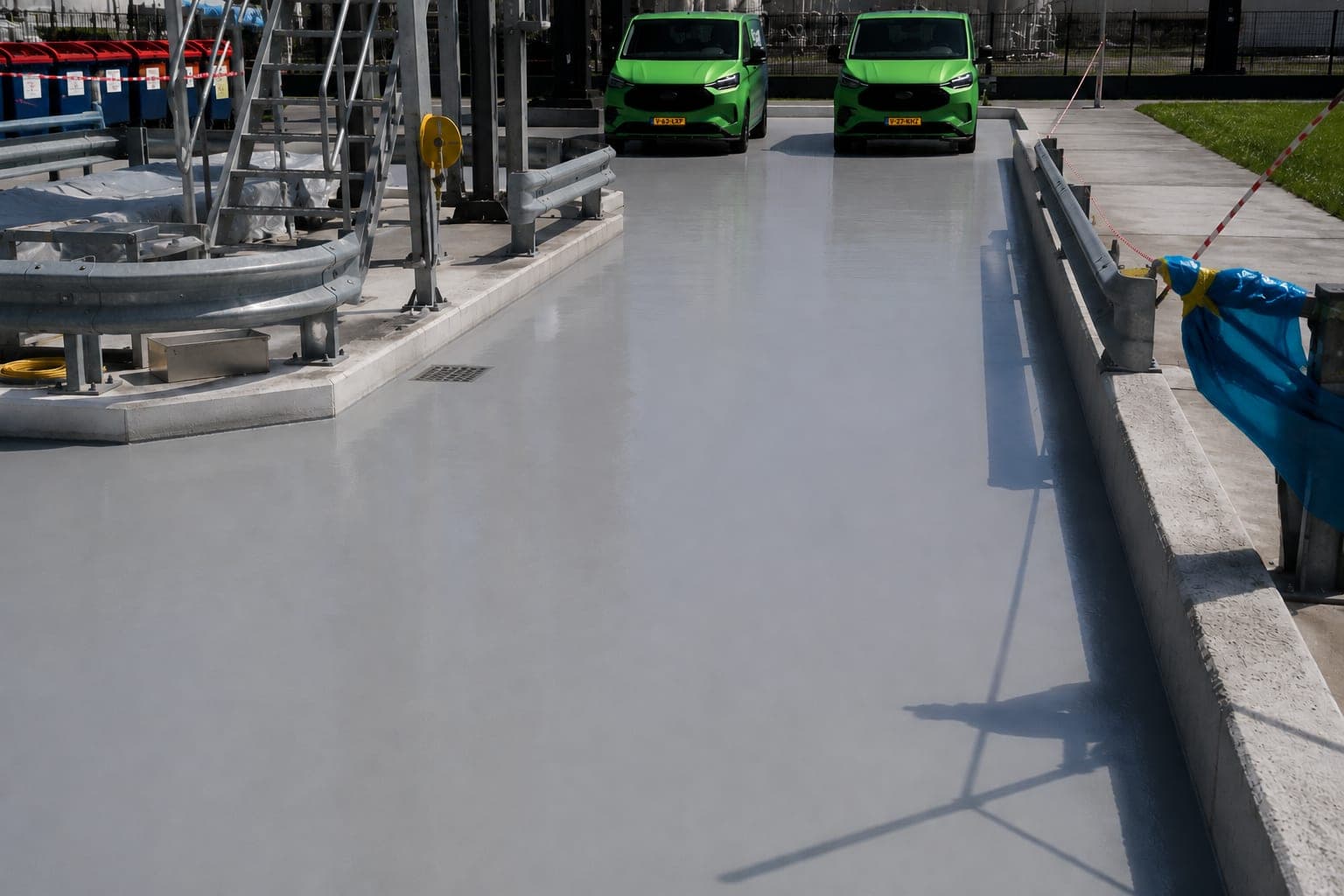 Industrieterrein met een gladde lichtgrijze gecoate vloer en een afvoerput, met vangrails, een stalen trap en twee groene bestelbussen op de achtergrond.