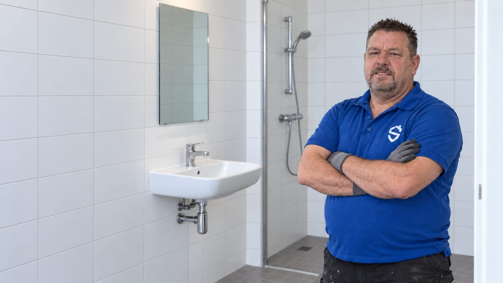 Man in een blauwe polo met het logo van Stevig Kitwerk staat met gekruiste armen en werkhandschoenen aan in een pas opgeleverde badkamer.