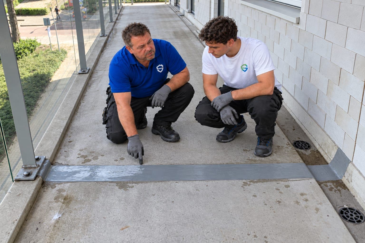 Twee mannen in werkkleding met het logo van Stevig Kitwerk zitten gehurkt bij een dilatatievoeg op een galerij. De oudste wijst naar de voeg, de jongste kijkt mee.