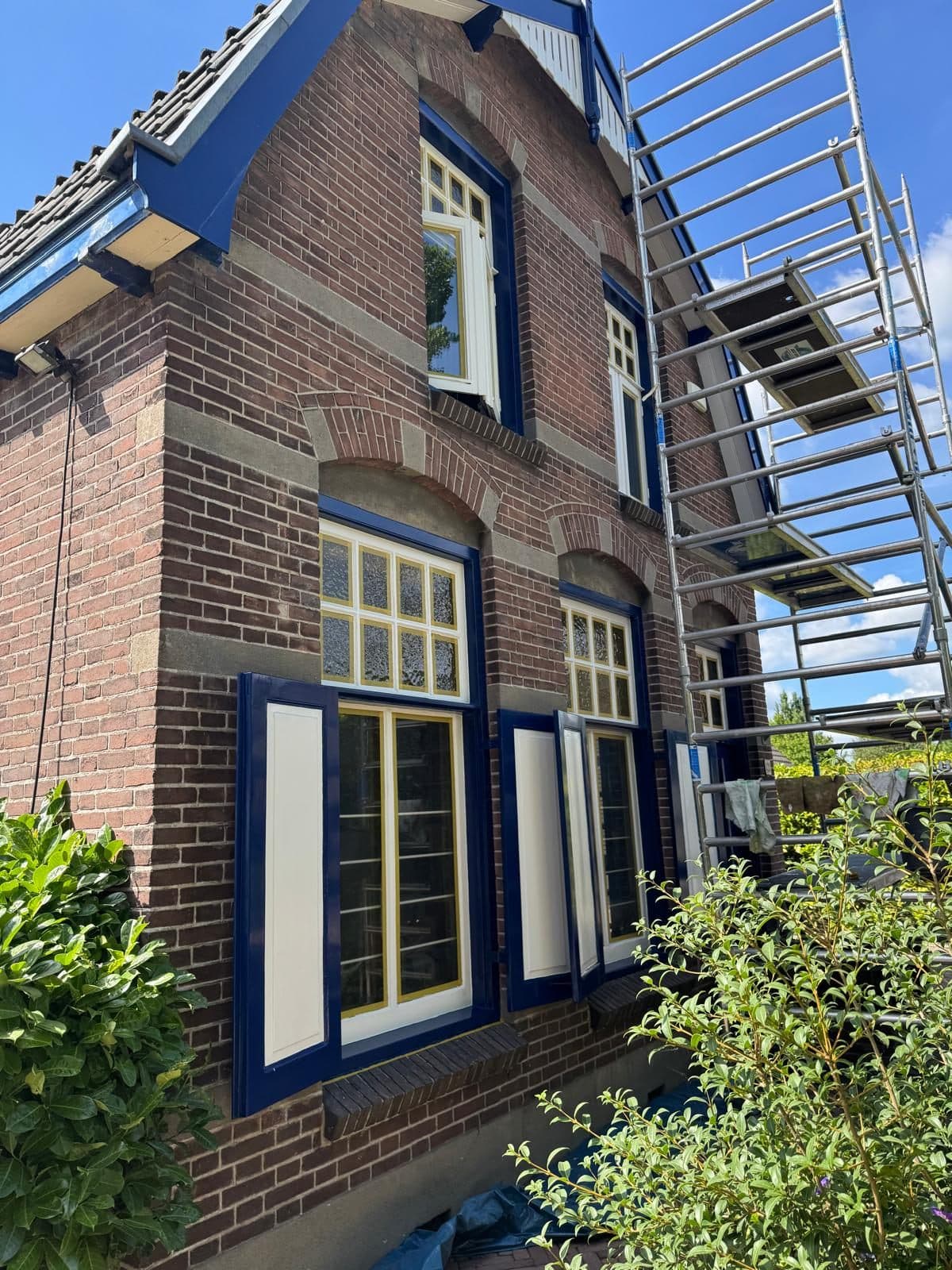 Bakstenen gevel met blauw geschilderde kozijnen en luiken, met een rolsteiger ernaast.