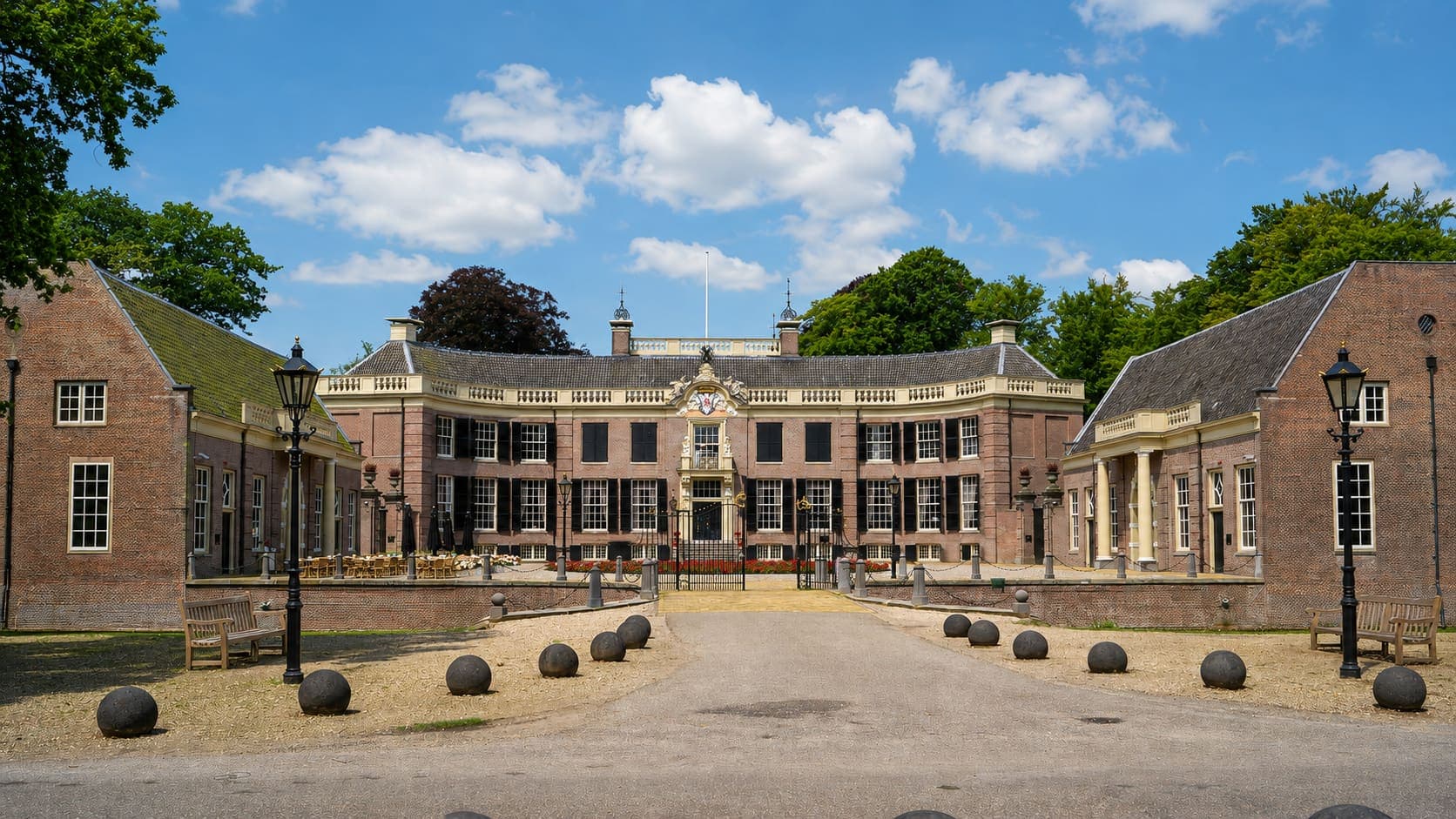 Symmetrisch landhuis van baksteen met een licht gepleisterde middenpartij, zwarte luiken en twee lage zijvleugels, met ervoor een grindoprit met stenen bollen, gietijzeren lantaarns en banken.