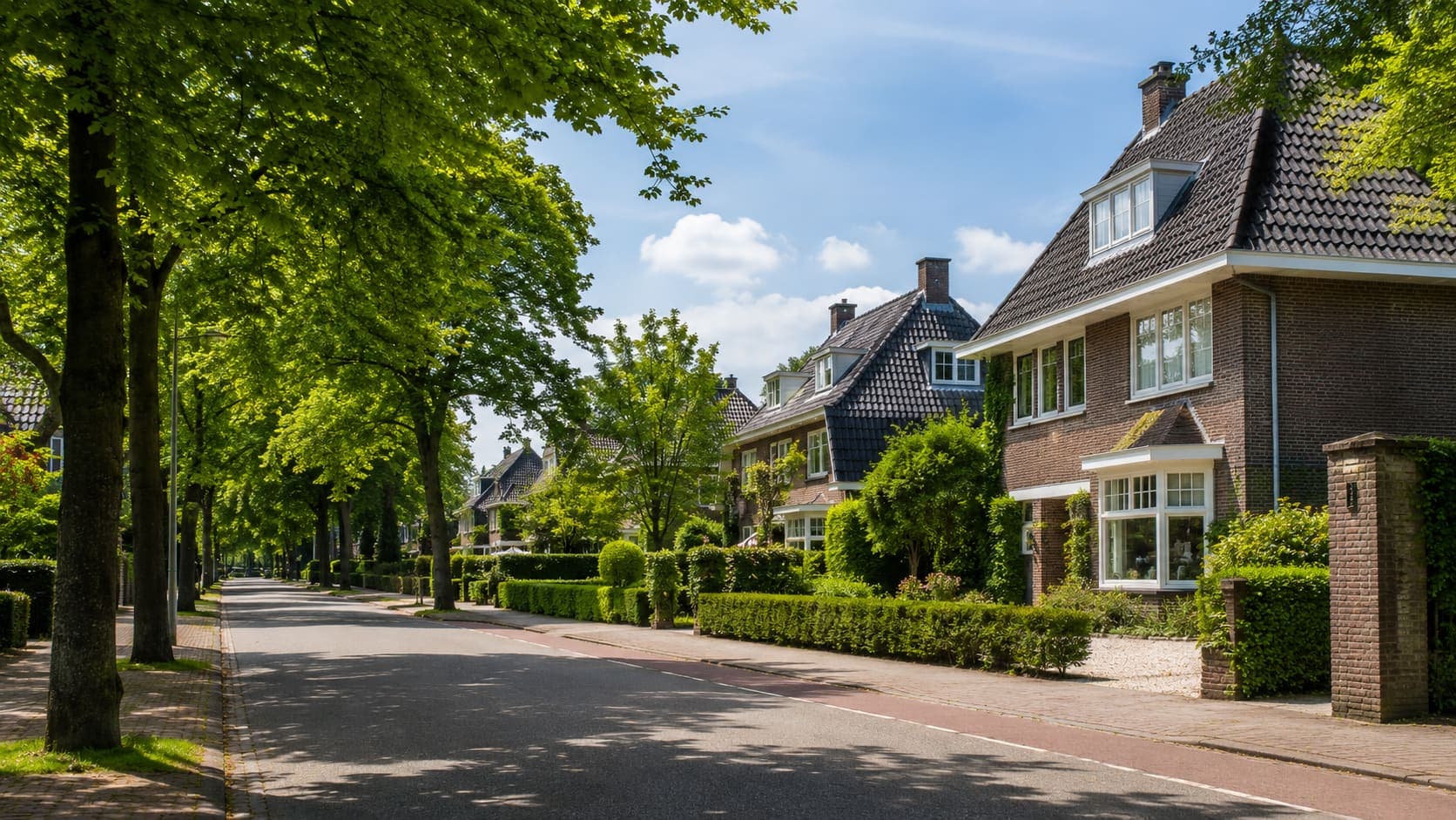 Villastraat met grote bakstenen huizen onder steile pannendaken, witte erkers en dakkapellen, achter geschoren hagen en een rij volgroeide bomen langs de weg.