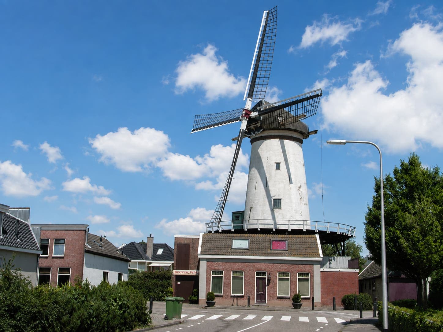Witte ronde stellingmolen met vier wieken boven een bakstenen onderbouw met winkelpui, aan een kruising met zebrapad, met woonhuizen en een boom ernaast.