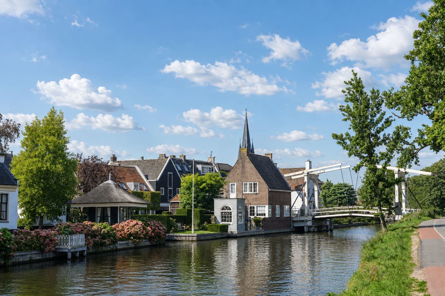 Vaart met langs de linkeroever huizen met pannendaken, een rieten theekoepel en bloeiende hortensia's, een kerktoren met spitse naald erachter, en rechts een witte ophaalbrug.