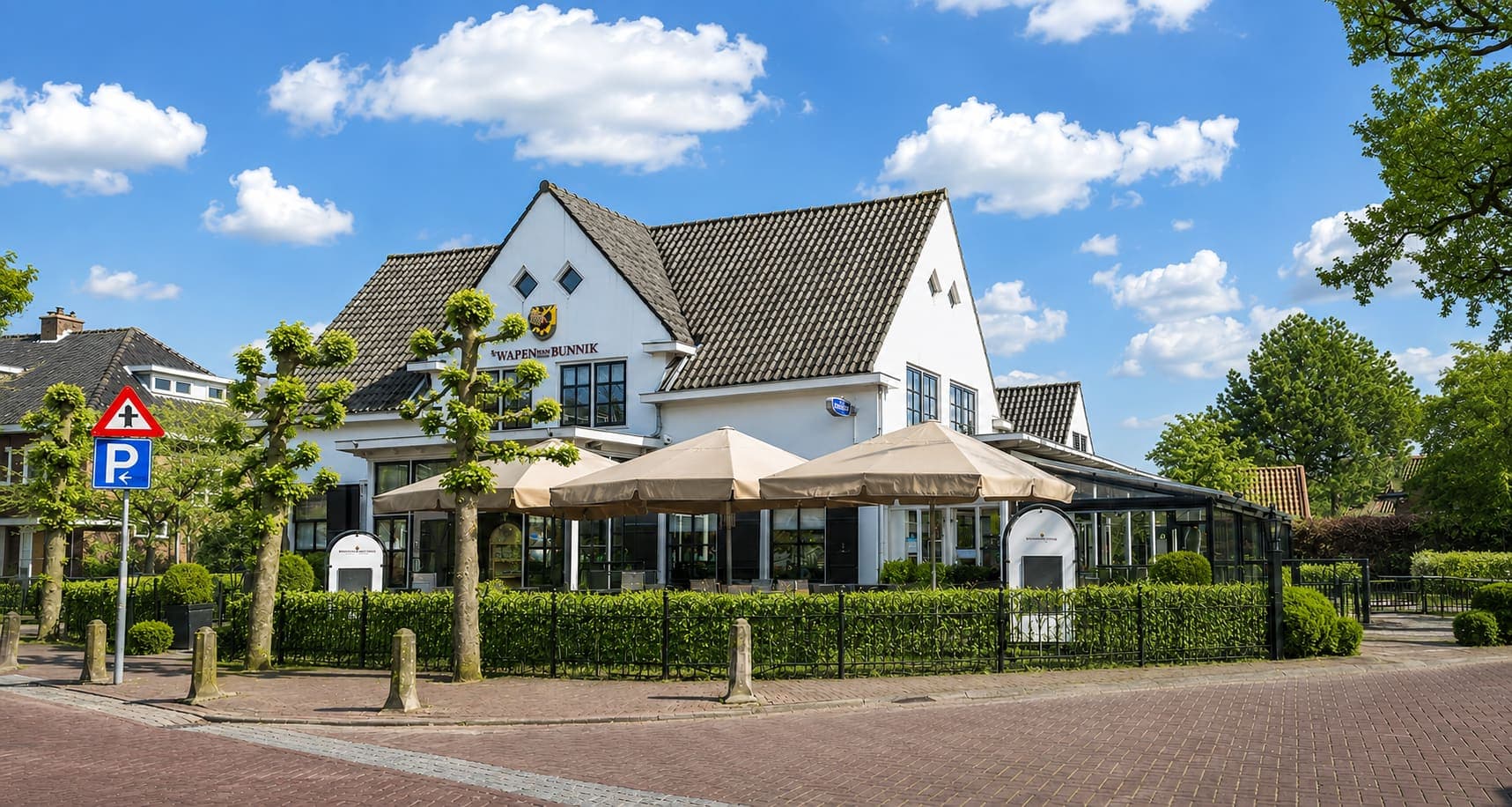 Wit gepleisterd pand met een steil pannendak en puntgevel, met ervoor een terras onder parasols, een lage haag en geknotte bomen langs de straat.