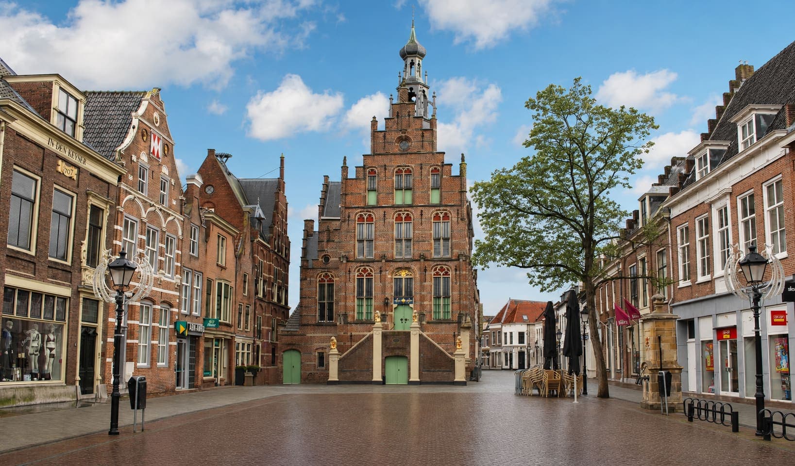 Stadhuis met een hoge trapgevel en een opengewerkte torenspits aan een geplaveid plein, met aan weerszijden bakstenen panden met winkelpuien en een boom rechts.