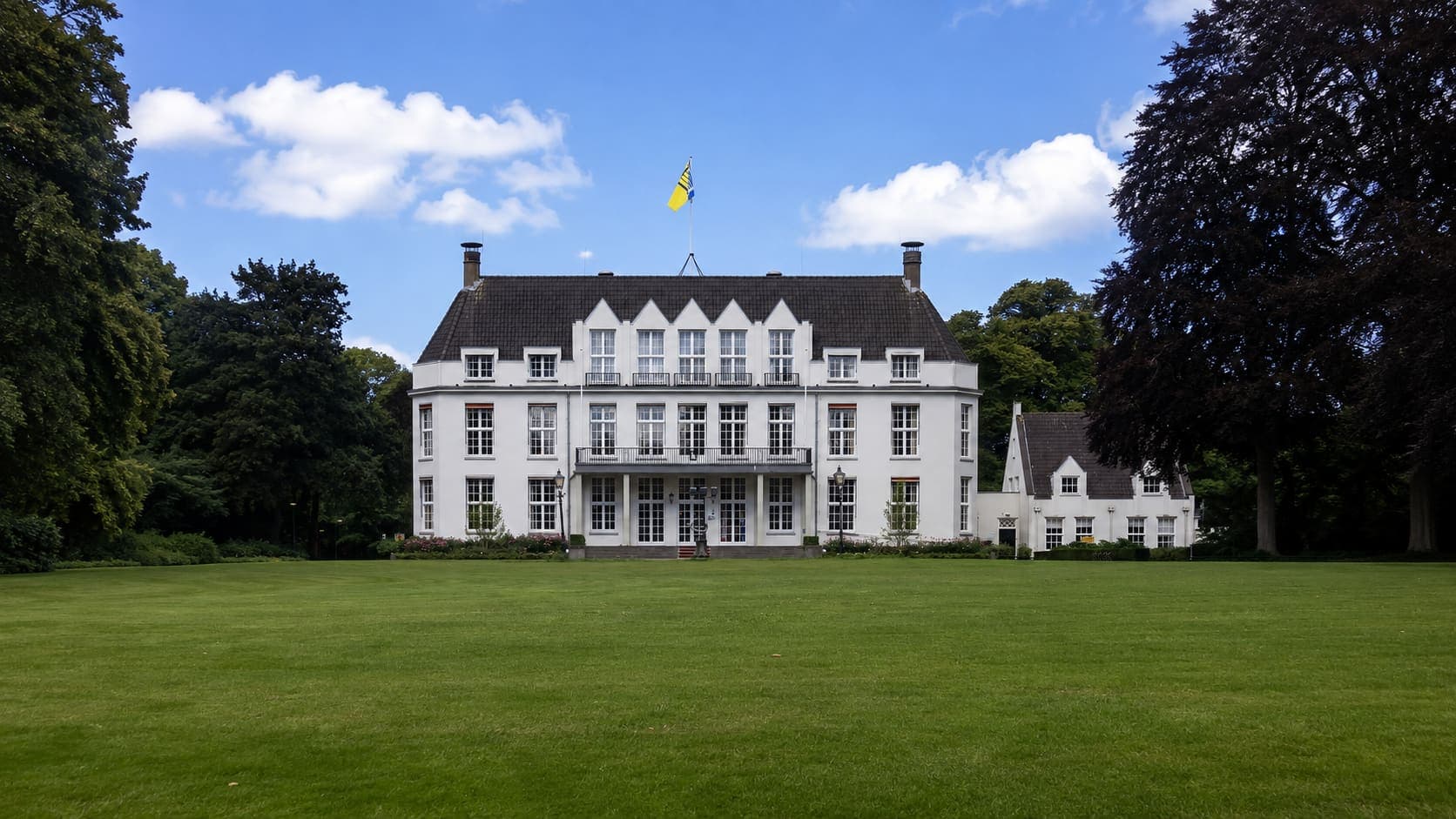 Wit landhuis met een leien schilddak, dakkapellen en een vlaggenmast, met een breed gazon ervoor en hoge bomen aan weerszijden.