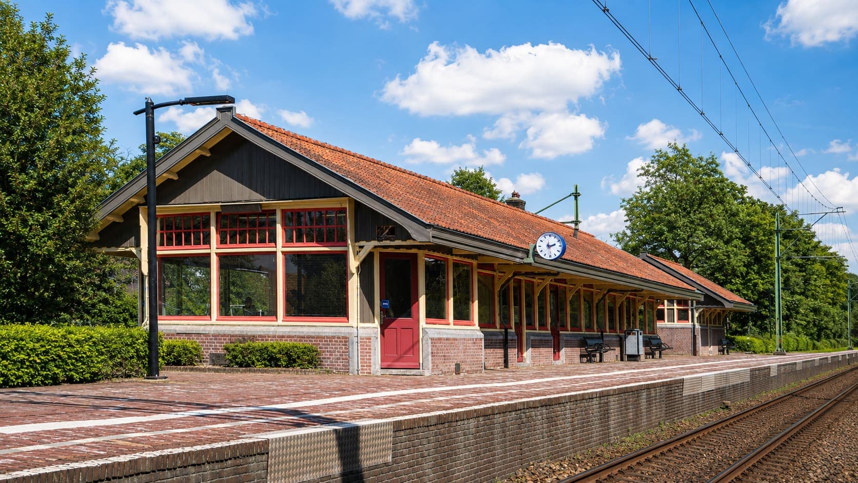 Laag stationsgebouw met een oranje pannendak, rood-geel geverfd houtwerk, een grote stationsklok en een overkapping langs het perron, met de rails ervoor.