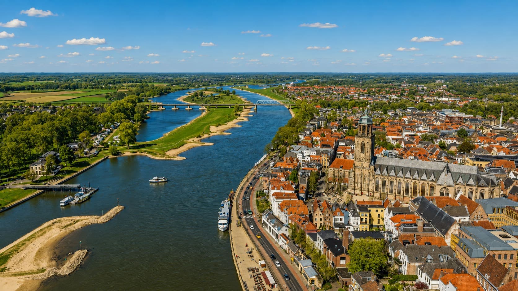 Uitzicht over een rivier met uiterwaarden, met op de rechteroever een stad en een grote gotische kerk.