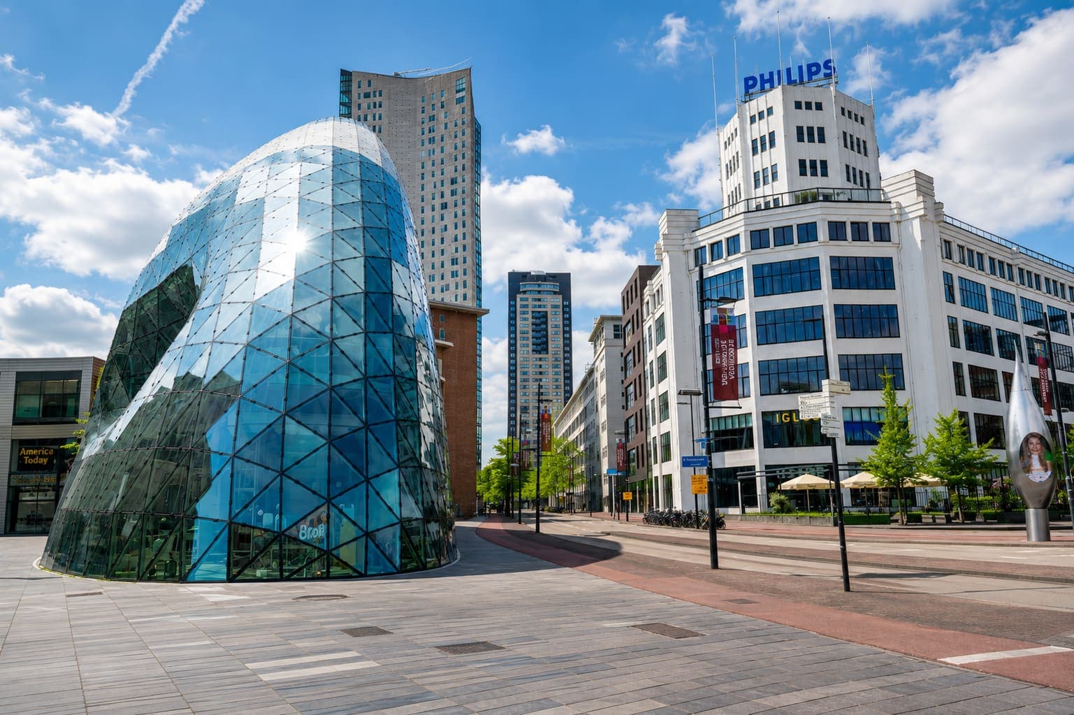Plein met een glazen gebouw in de vorm van een druppel, en daarachter hoogbouw en het witte Philips-gebouw met lichtreclame op het dak.
