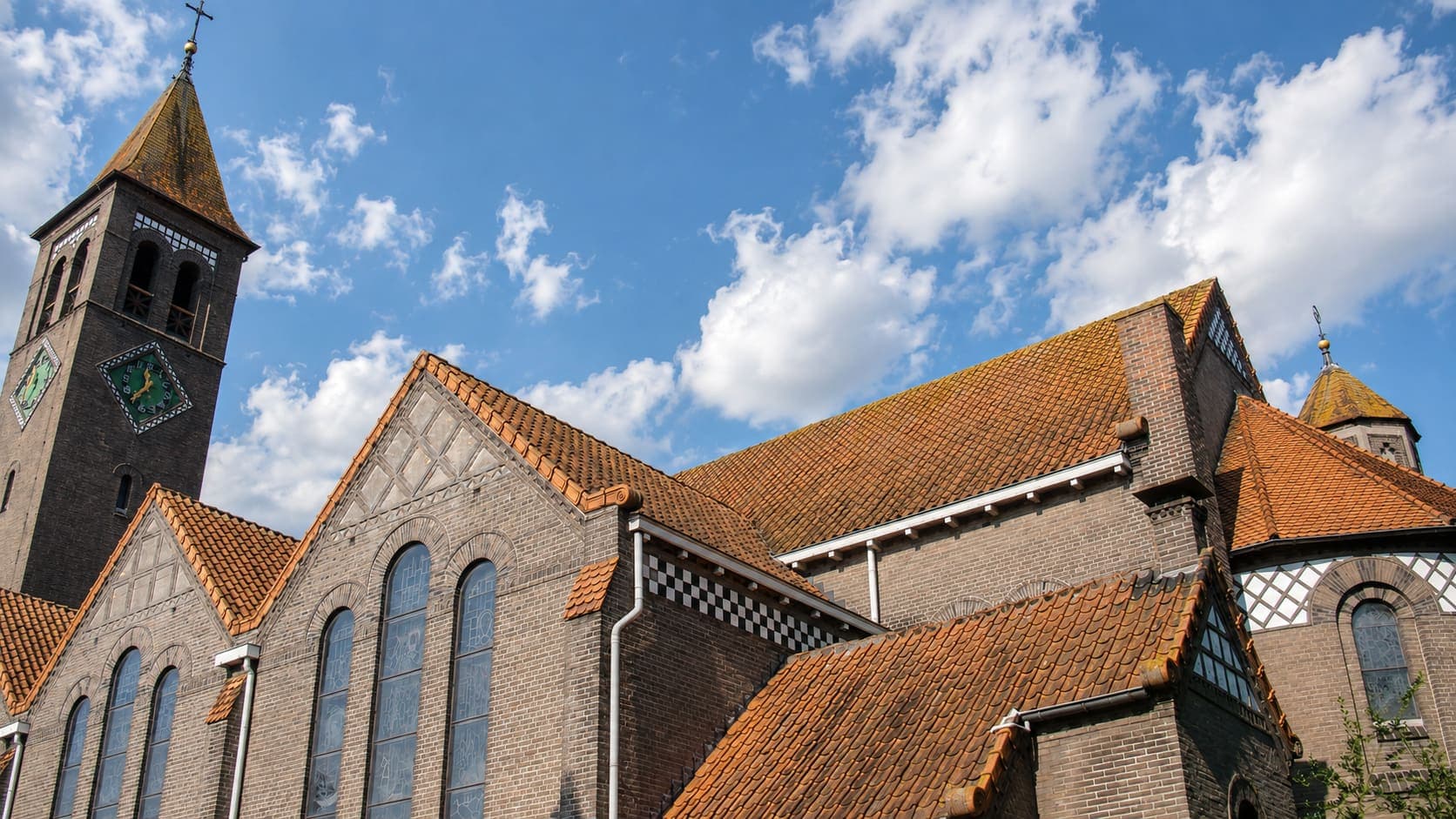 Grote bakstenen kerk van onderaf gezien, met een donkere klokkentoren met een groene wijzerplaat, oranje pannendaken en hoge boogramen met glas-in-lood.