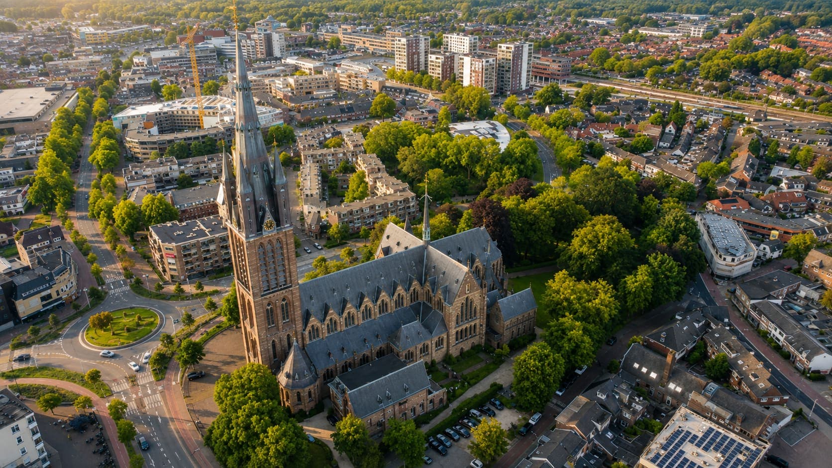 Luchtfoto over Hilversum met een grote bakstenen kerk met een hoge spitse toren, omringd door groen, woonstraten en een rotonde.