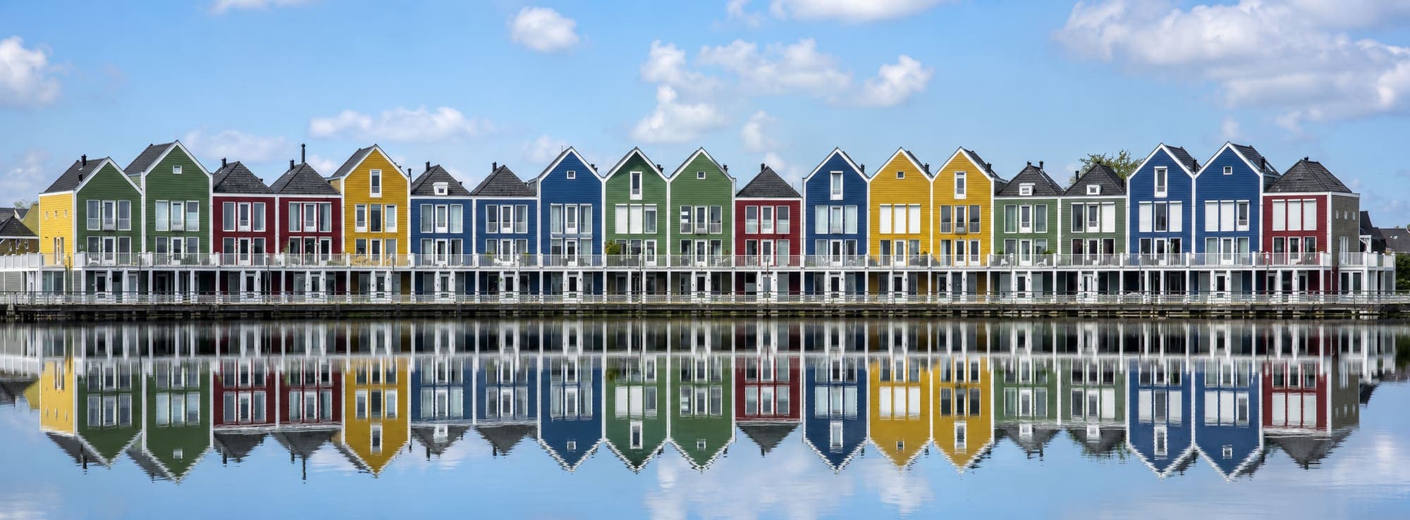 Rij smalle huizen met houten gevels in geel, rood, blauw en groen langs het water, met puntgevels en witte balkons, weerspiegeld in de vlakke waterspiegel ervoor.