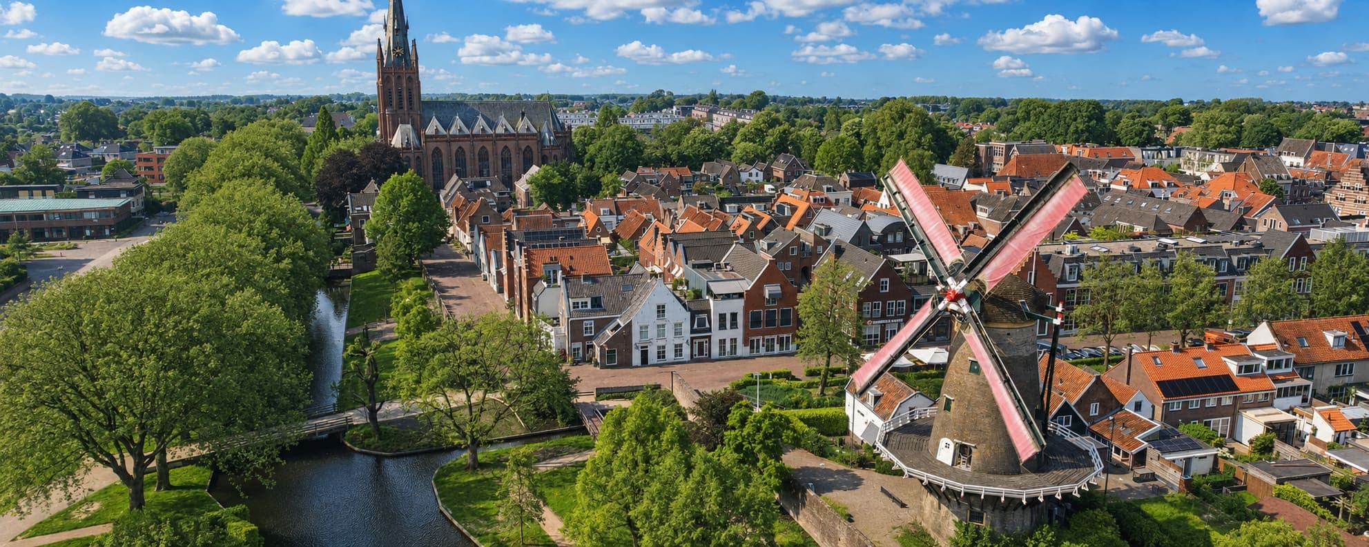 Luchtfoto van een oude stadskern met een hoge bakstenen kerktoren, een stellingmolen met vier roeden, rijen huizen met rode pannendaken en een gracht met bomen erlangs.
