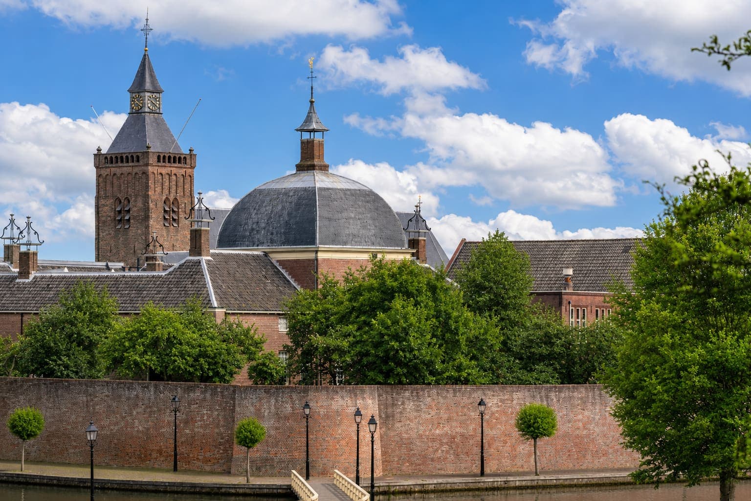 Bakstenen kerktoren met een leien spits en wijzerplaat naast een achthoekige koepelkerk met loden dak, boven een bakstenen stadsmuur met lantaarns en leibomen langs het water.