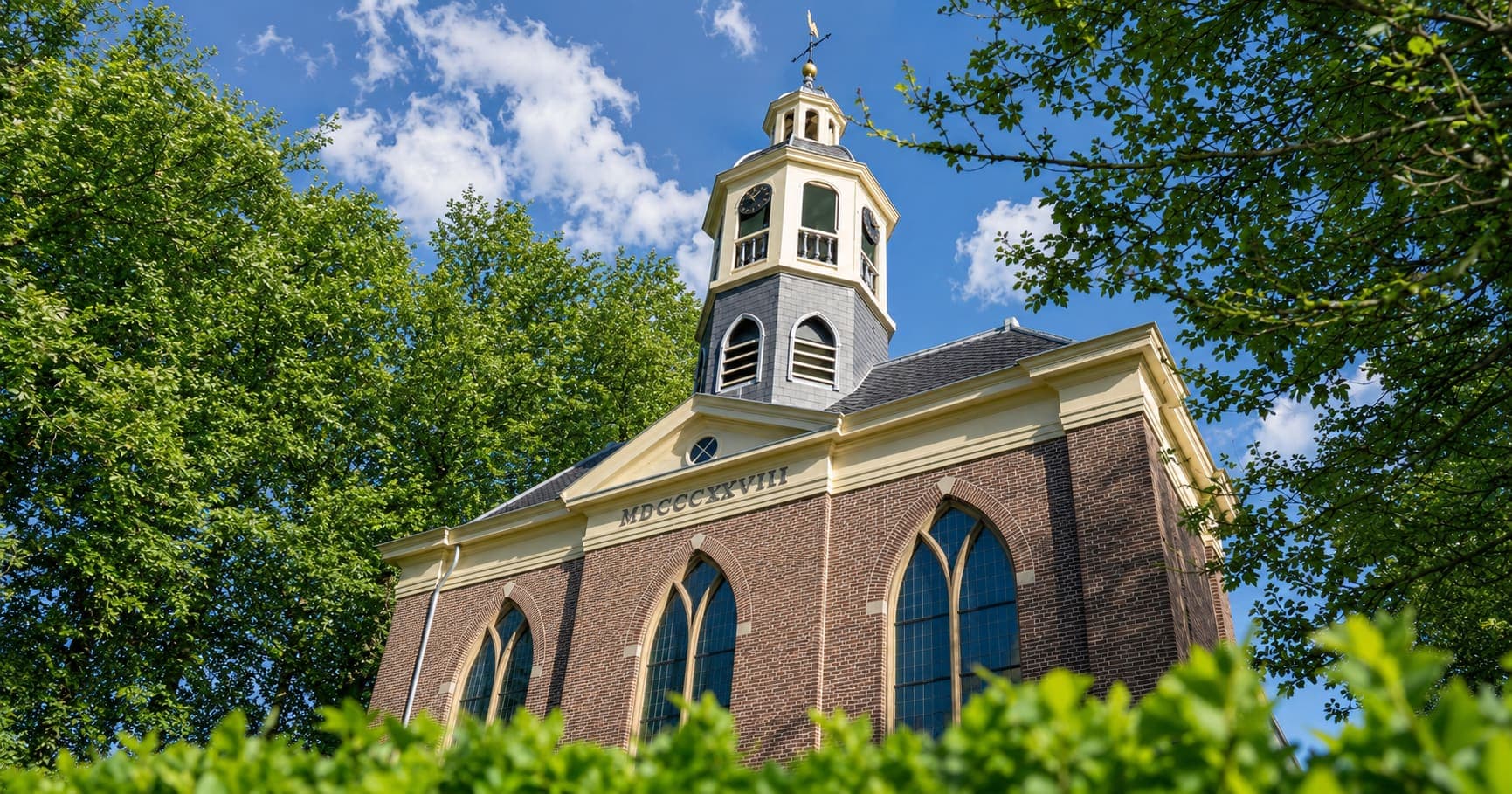 Bakstenen dorpskerk van onderaf gezien, met spitsboogramen, een geel geschilderd fronton met een jaartal in Romeinse cijfers en een achtkantige klokkentoren met wijzerplaat en weerhaan, omzoomd door bomen.