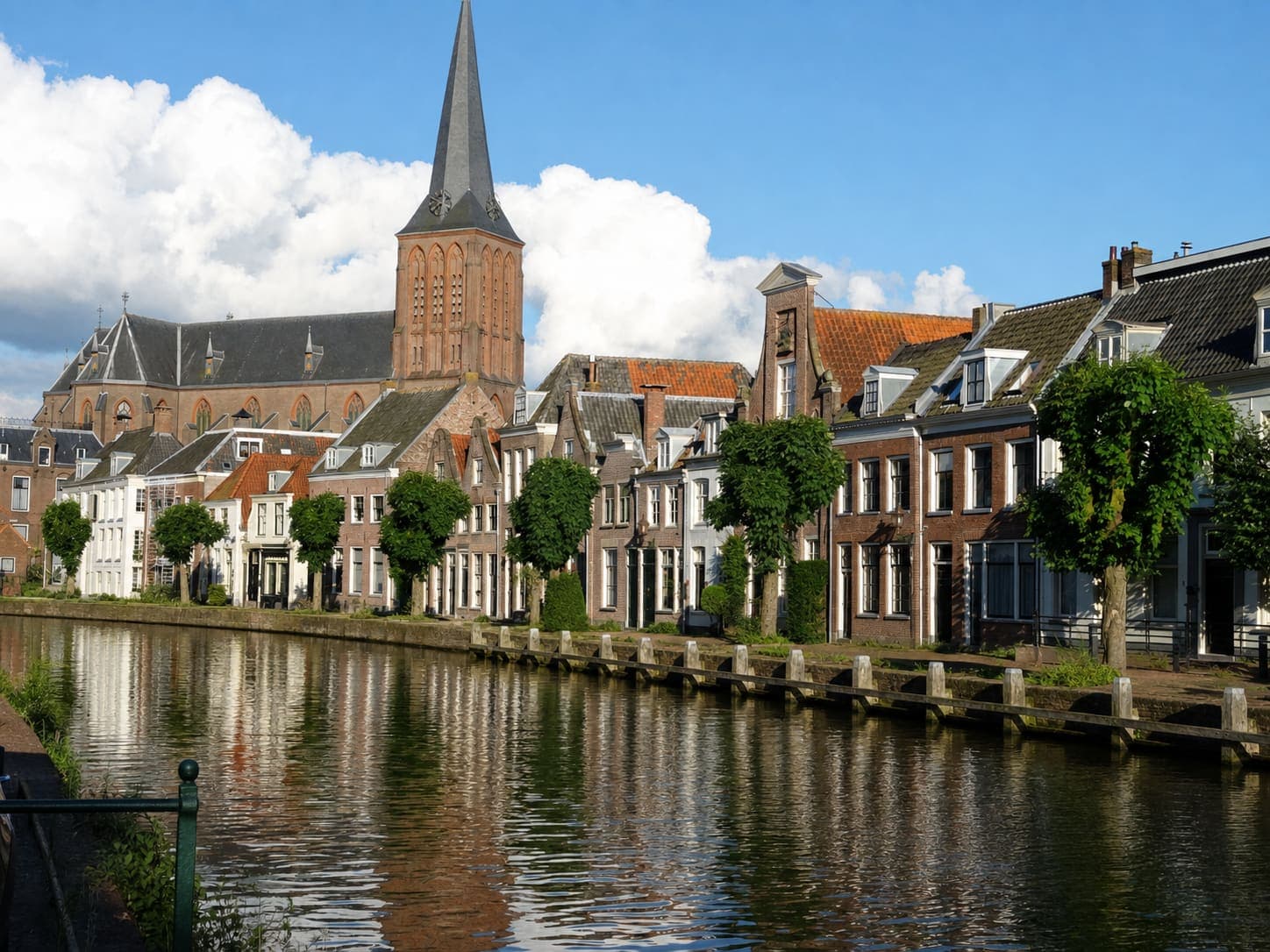Historische panden langs de Vecht met een bakstenen kerktoren met hoge spits erboven, geschoren bomen langs de kade en de gevels weerspiegeld in het water.