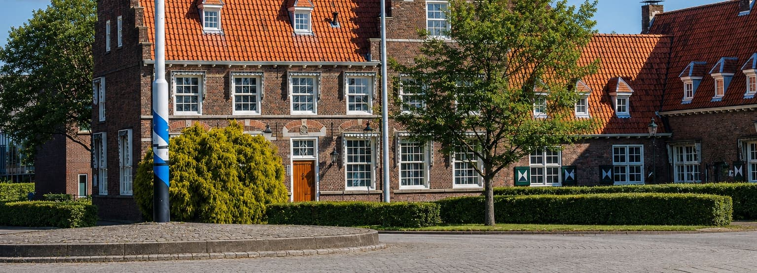 Langgerekt bakstenen pand met een oranje pannendak, dakkapellen en witte schuiframen, met ervoor een lage haag, een jonge boom en een geplaveid plein.