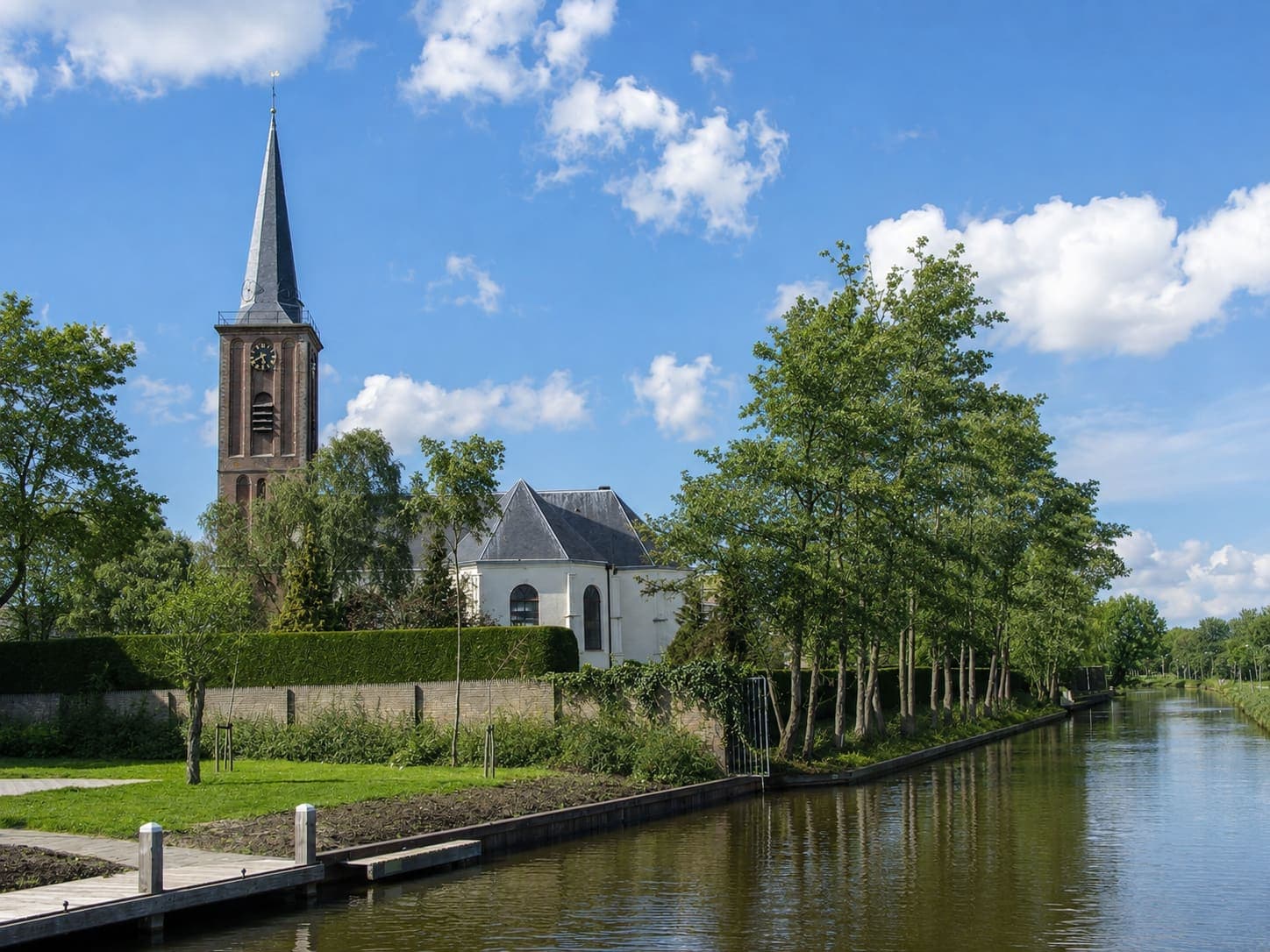 Bakstenen kerktoren met een hoge leien spits, een wijzerplaat en galmborden, naast een witgepleisterde kerk met leien daken, achter een geschoren haag langs een vaart met beschoeide oever.