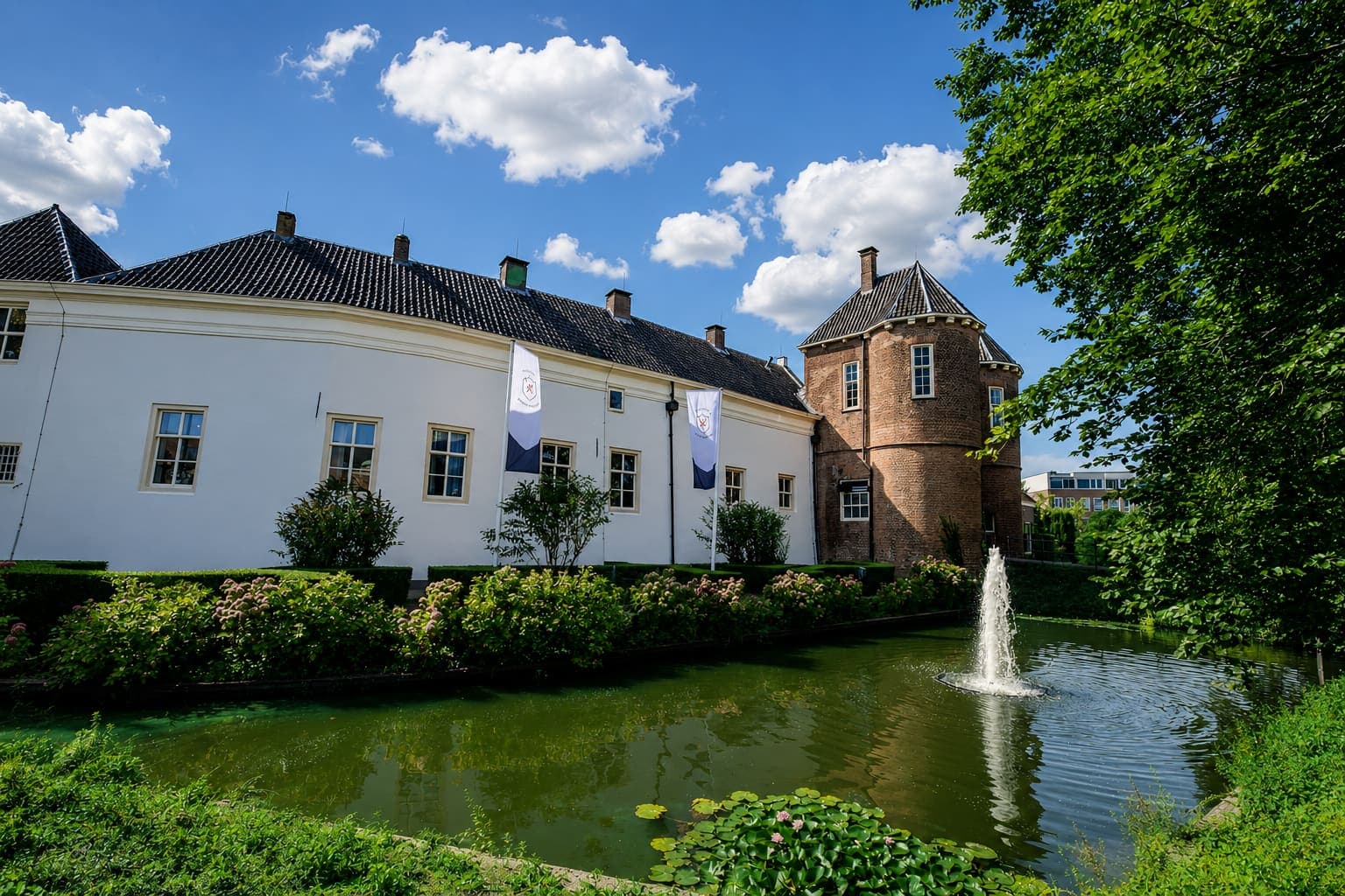 Wit gepleisterd gebouw met een zwart pannendak en een ronde bakstenen toren, aan een gracht met een fontein, met een lage haag en bloeiende struiken langs het water en twee vlaggen aan masten tegen de gevel.