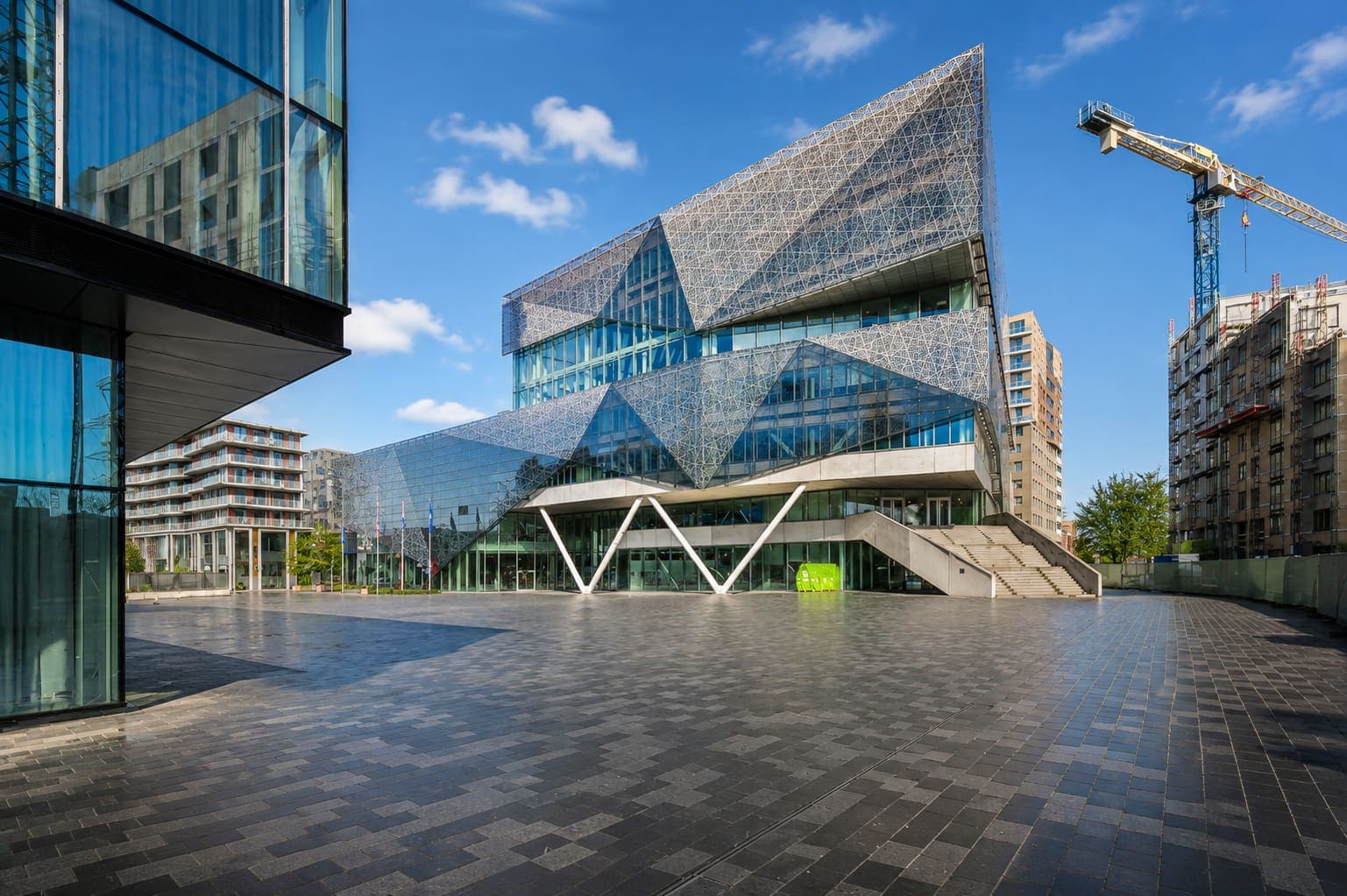 Gebouw met hoekige glasgevels en een wit netwerk eroverheen, aan een geplaveid plein, met een bouwkraan erachter.