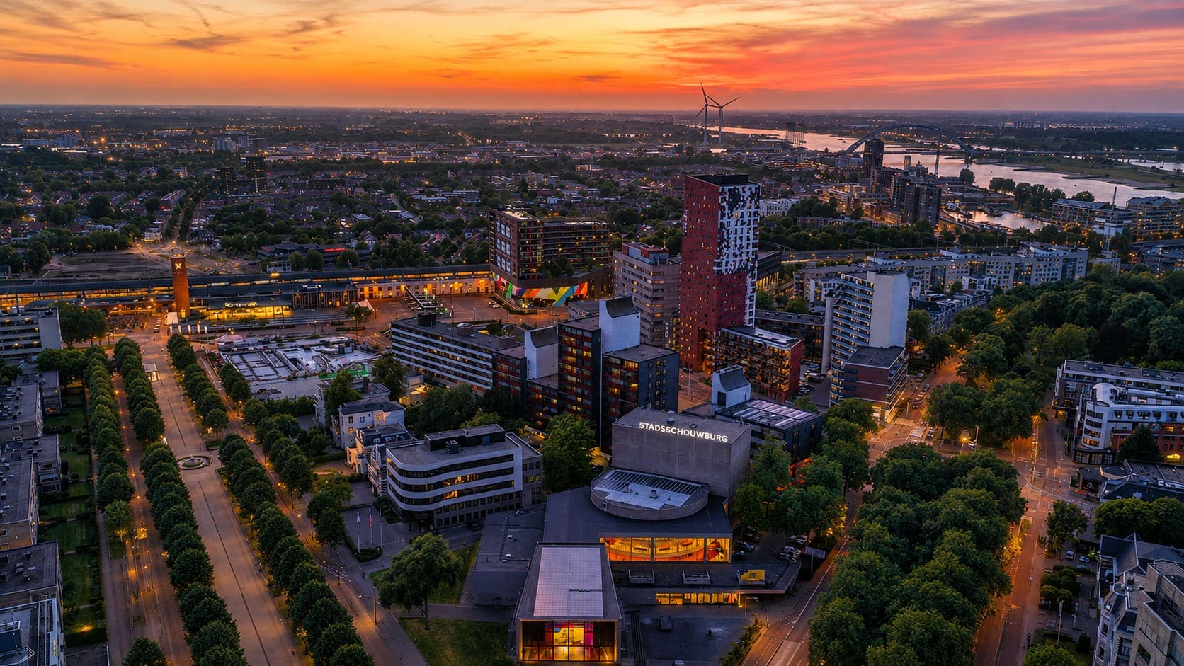Uitzicht bij zonsondergang over een stad, met een schouwburg op de voorgrond, hoogbouw erachter en een rivier met een boogbrug aan de horizon.