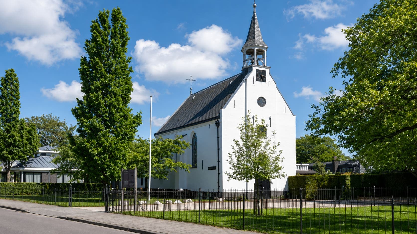 Witte dorpskerk met een leien zadeldak en een open klokkentoren met spits, achter een zwart hek, met bomen en een grasveld ervoor.