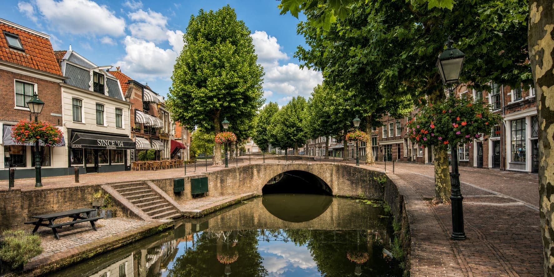 Gemetselde boogbrug over een gracht in een oude stadskern, met aan weerszijden bakstenen panden met winkelpuien en zonneweringen, bloembakken aan gietijzeren lantaarns en een rij platanen langs de kade.