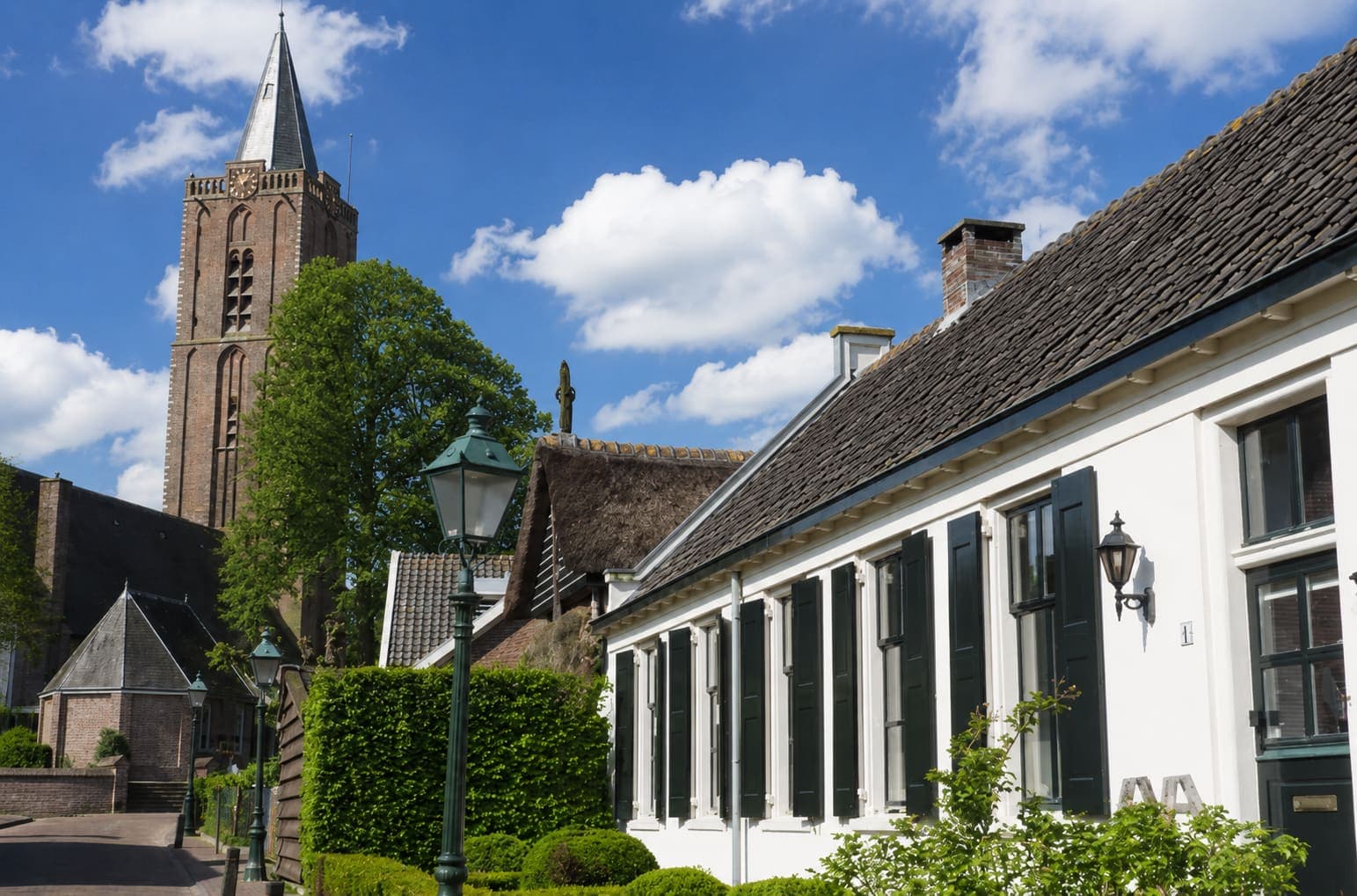 Bakstenen kerktoren met een leien spits en wijzerplaat boven de bomen, met op de voorgrond een wit gepleisterd huis met donkergroene luiken, een rieten dak ernaast en een gietijzeren lantaarn bij een geschoren haag.