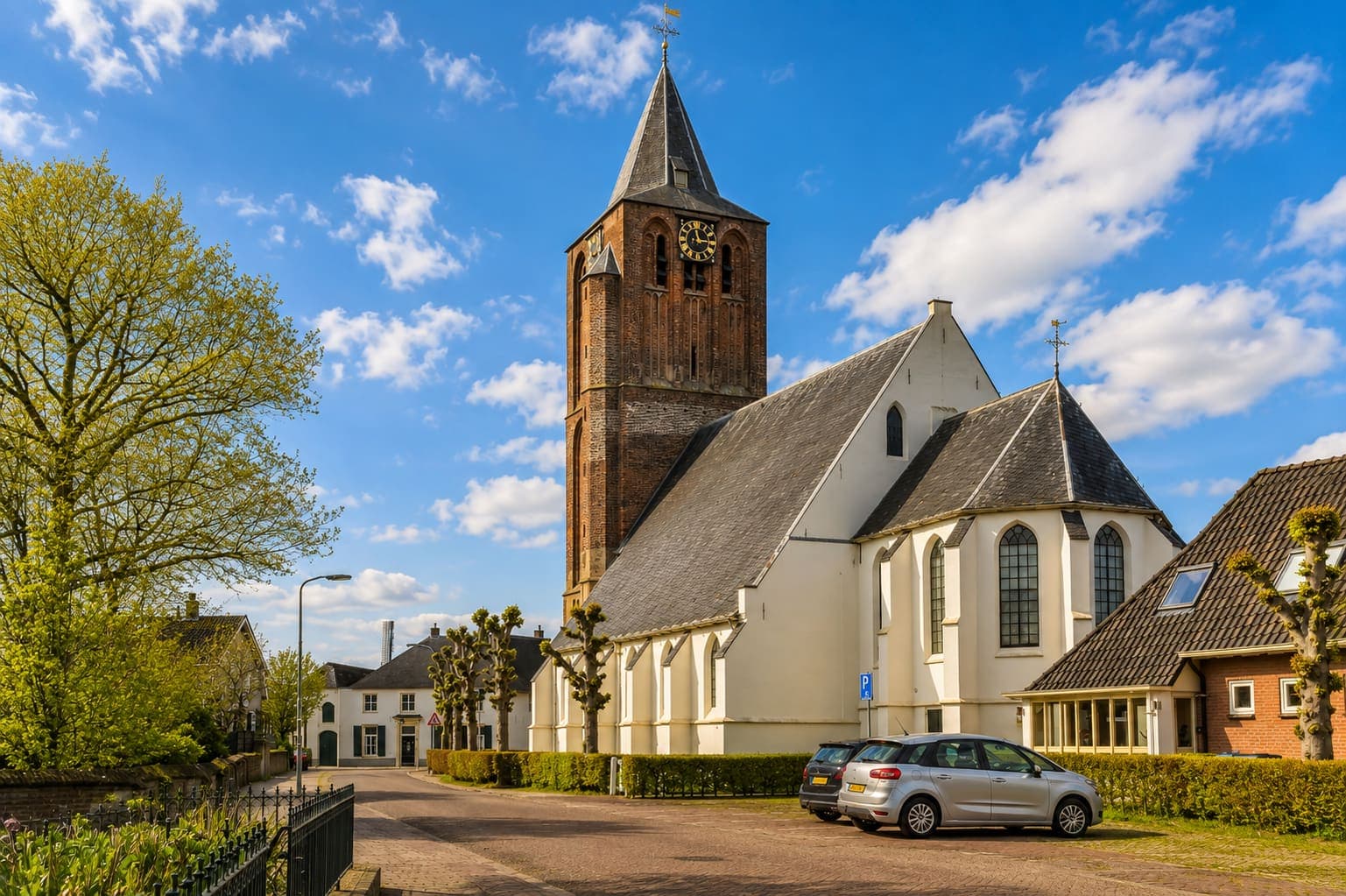 Witgepleisterde kerk met een bakstenen toren met uurwerk, aan een straat met geknotte bomen.