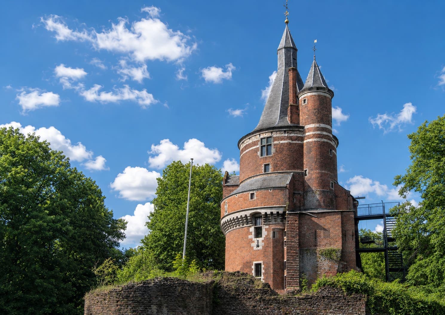 Ronde bakstenen kasteeltoren met twee leien spitsen, kruisvensters en een uitkragende weergang op consoles, met een begroeide ruïnemuur ervoor en hoge bomen aan weerszijden.
