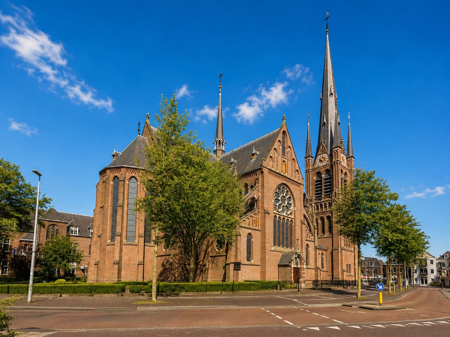 Grote bakstenen kerk met een hoge spitse toren en een roosvenster, aan een kruispunt.