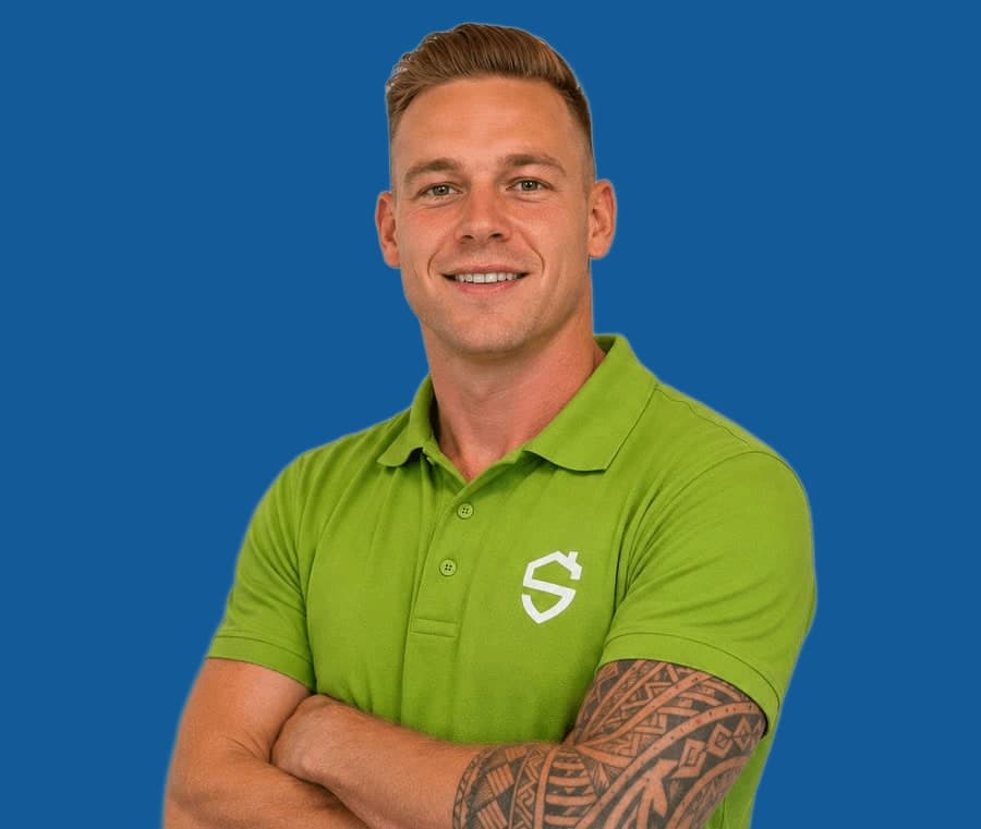 Man met kort blond haar en een getatoeëerde onderarm in een groene bedrijfspolo, armen over elkaar, op een effen blauwe achtergrond.
