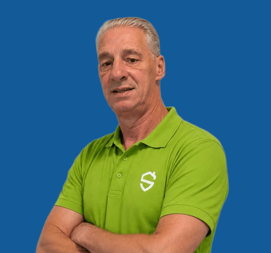 Man met grijs haar in een groene bedrijfspolo, armen over elkaar, op een effen blauwe achtergrond.